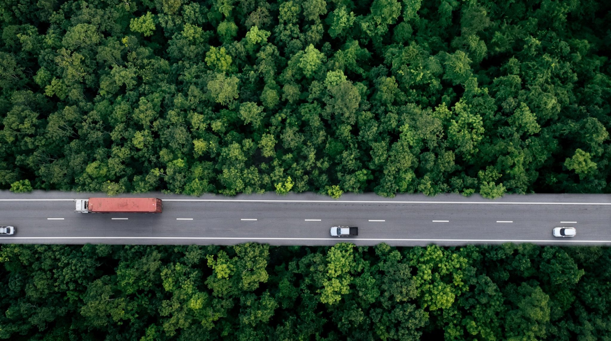 Aerial Top View Of Car And Truck Driving On Highway Road In Green Forest. Sustainable Transport. Drone View Of Hydrogen Energy Truck And Electric Vehicle Driving On Asphalt Road Through Green Forest.